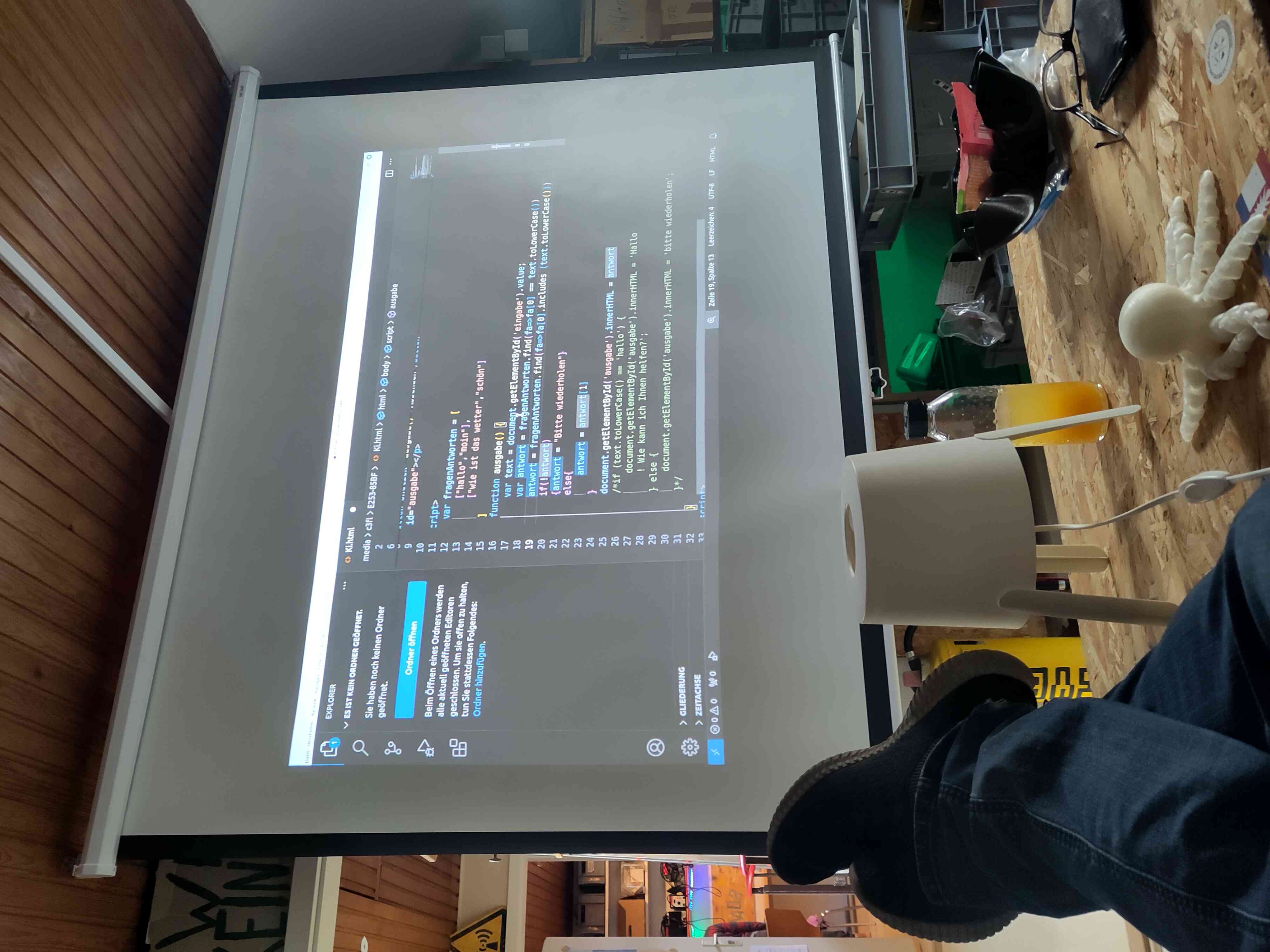Auf einem Beamer ist Programm Code in sehr klein zu sehen. Es handelt sich um eine KI.html. im Vordergrund sind Füße auf einem Tisch zu sehen